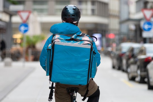 Eine Fahrrad fahrende Person ist von hinten zu sehen. Die Person trägt einen dunklen Helm, sowie eine Jacke und einen Lieferdienst-Rucksack im gleichen hellblauen Farbton. 