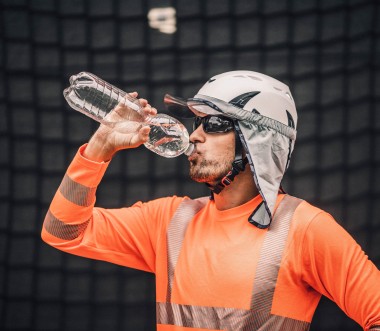 Ein Arbeitnehmer trägt Kleidung zum Schutz gegen Hitze und trinkt Wasser aus einer Flasche. Ein Arbeitnehmer trägt Kleidung zum Schutz gegen Hitze und trinkt Wasser aus einer Flasche.