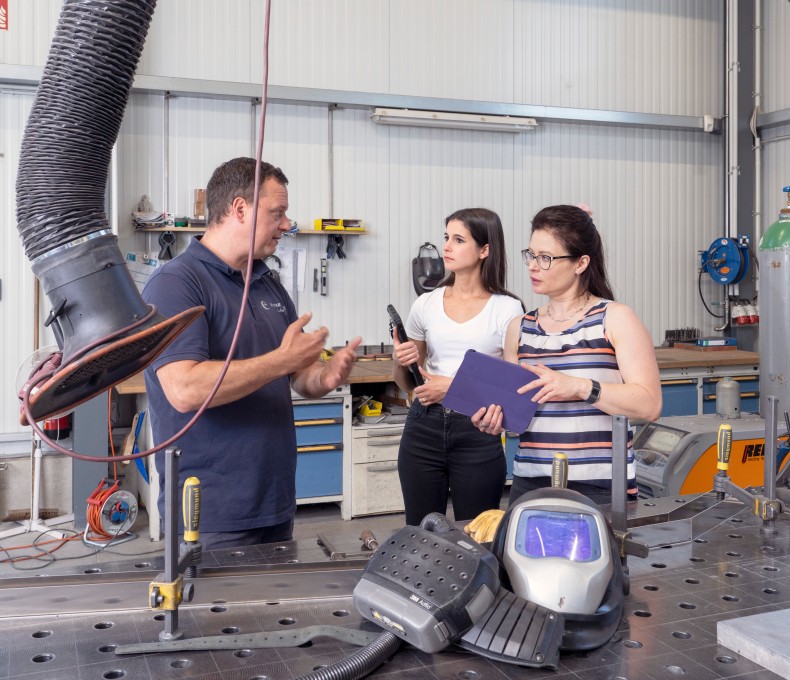 In einer Werkstatt mit metallverarbeitender Ausstattung stehen ein Mann und zwei Frauen im Gespräch. Der Mann trägt ein dunkelblaues Poloshirt und erklärt gestikulierend etwas. Die eine Frau hält ein Klemmbrett, die andere ein Werkzeug. Im Vordergrund liegt ein Schweißhelm auf einem großen Metallarbeitstisch, an dem Absaugvorrichtungen und weitere Werkzeuge angebracht sind. Im Hintergrund sind Werkbänke, Gasflaschen und weitere Arbeitsmittel sichtbar.
In einer Werkstatt mit metallverarbeitender Ausstattung stehen ein Mann und zwei Frauen im Gespräch. Der Mann trägt ein dunkelblaues Poloshirt und erklärt gestikulierend etwas. Die eine Frau hält ein Klemmbrett, die andere ein Werkzeug. Im Vordergrund liegt ein Schweißhelm auf einem großen Metallarbeitstisch, an dem Absaugvorrichtungen und weitere Werkzeuge angebracht sind. Im Hintergrund sind Werkbänke, Gasflaschen und weitere Arbeitsmittel sichtbar.