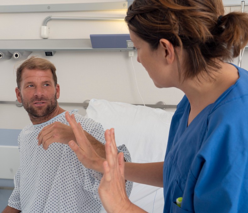 Ein Mann in Krankenhauskleidung sitzt aufrecht im Bett und schaut eine Pflegekraft an, die ihm gegenübersitzt und mit erhobenen Händen eine abwehrende Geste macht. Die Situation wirkt angespannt. Ein Mann in Krankenhauskleidung sitzt aufrecht im Bett und schaut eine Pflegekraft an, die ihm gegenübersitzt und mit erhobenen Händen eine abwehrende Geste macht. Die Situation wirkt angespannt.