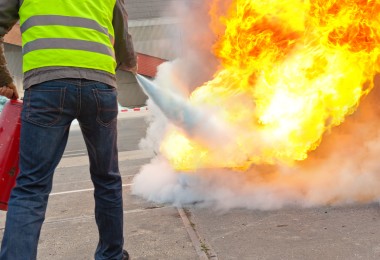 Eine Person mit Warnweste löscht bei einer Brandschutzübung im Freien mit einem Feuerlöscher einen großen, kontrollierten Flammenbrand.