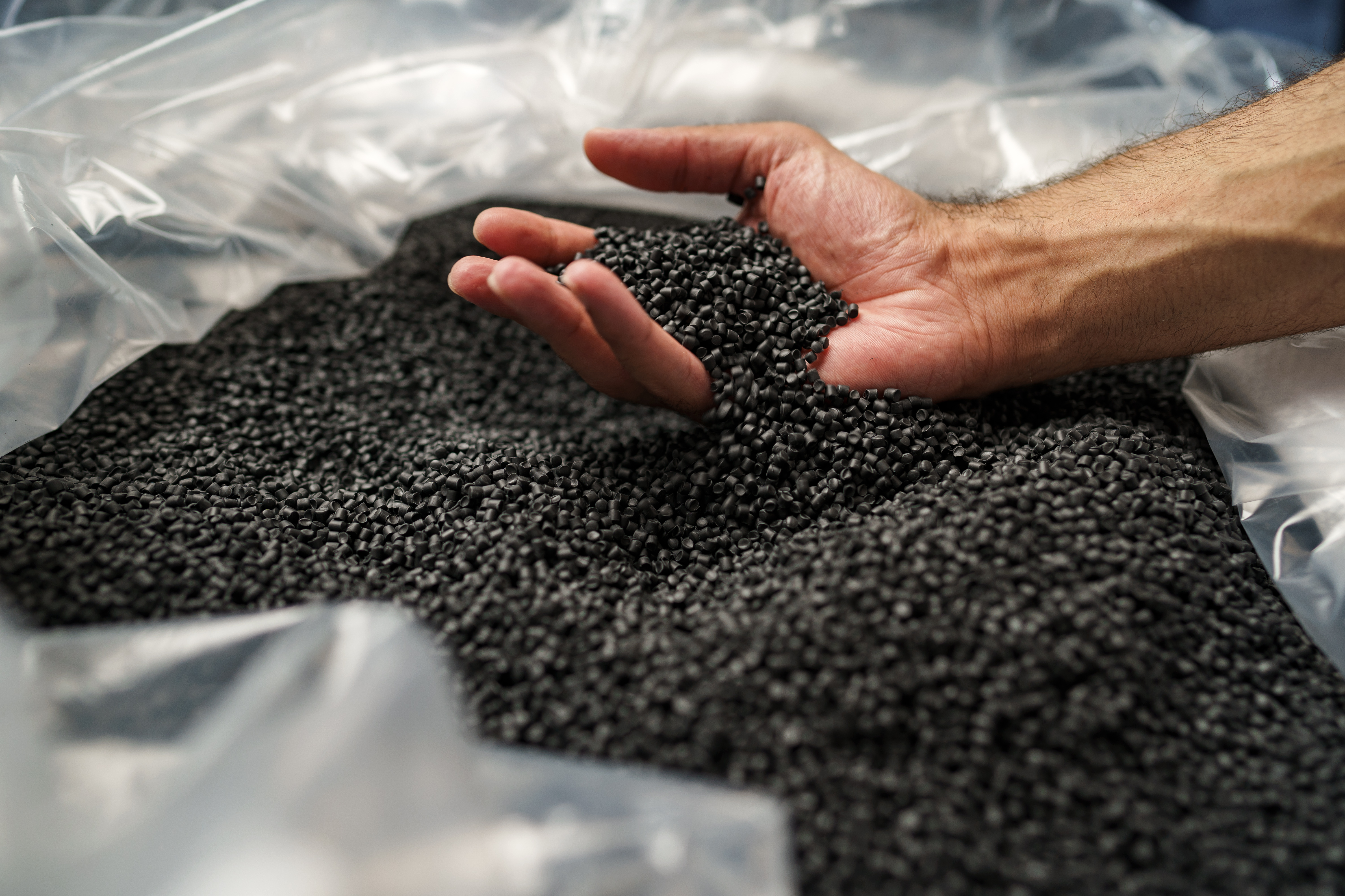 Eine Hand greift in einen großen Behälter mit schwarzen Kunststoffgranulaten und lässt einige der kleinen Körner durch die Finger gleiten. (Bild: fotofabrika / stock.adobe.com)