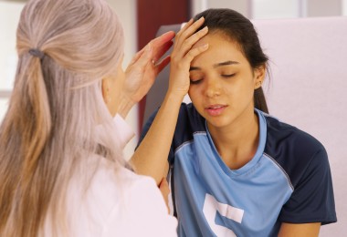 Ein junges Mädchen im blauen Trikot sitzt mit geschlossenen Augen und hält sich die Stirn. Eine zweite Frau untersucht vorsichtig ihren Kopf und stützt ihren Arm. (Bild: Mark Adams / stock.adobe.com)