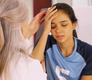 Ein junges Mädchen im blauen Trikot sitzt mit geschlossenen Augen und hält sich die Stirn. Eine zweite Frau untersucht vorsichtig ihren Kopf und stützt ihren Arm. (Bild: Mark Adams / stock.adobe.com)