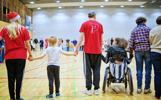 In einer Sporthalle sehen wir eine von links nach rechts die Rücken einer Reihe Menschen, die sich an den Händen halten. Sie sind unterschiedlich alt. Einige haben erkennbar eine Behinderung, andere nicht. Einige tragen T-Shirts des Vereins.