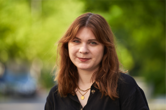 Portrait von Elena Everding, junge Frau mit eher rundem Gesicht und langen leicht gewellten Haaren vor Bäumen, die aber verschwommen sind.