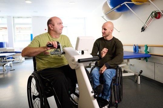 Jens sitzt rechts im Rollstuhl. Links von ihm sitzt ein anderer Mann im Rollstuhl und trainiert. Jens spricht zu ihm.
