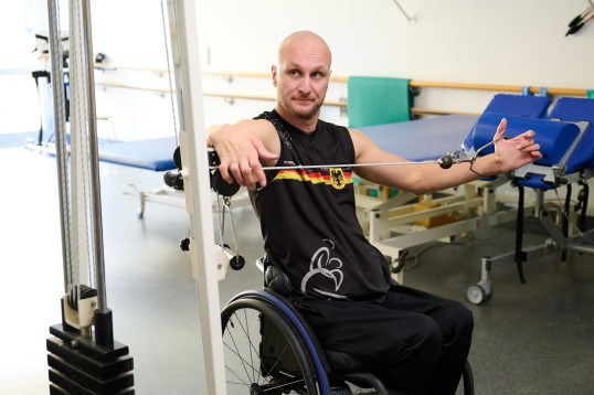 Jens Sauerbier beim Training. ImRollstuh sitzend zieht er seinen Arm seitwärts. Sein Arm ist mit einem Kabel verbunden, das ein Gewicht in die Höhe zieht.