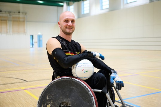 JensSauerbier von der Seite im Rugby-Rollstuhl sitzend. Er sieht direkt in die Kamera. Die Sporthalle, in der er sich befindet, ist sonst menschenleer.
