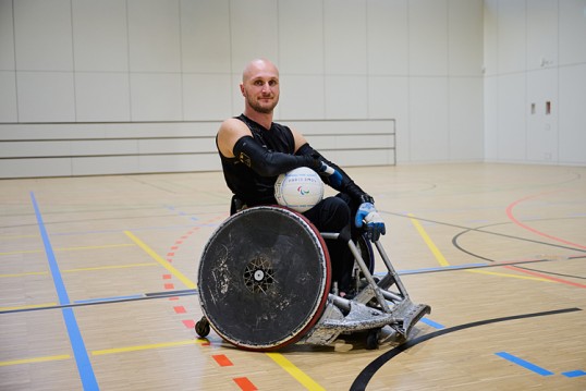 Jens Sauerbier im Rugby-Rollstuhl. Er befindet sich in einer sonst leeren Sporthalle und hält den Ball im Arm. Schlanker junger Mann, kurzer Vollbart, Glatze, schlankes Gesicht.